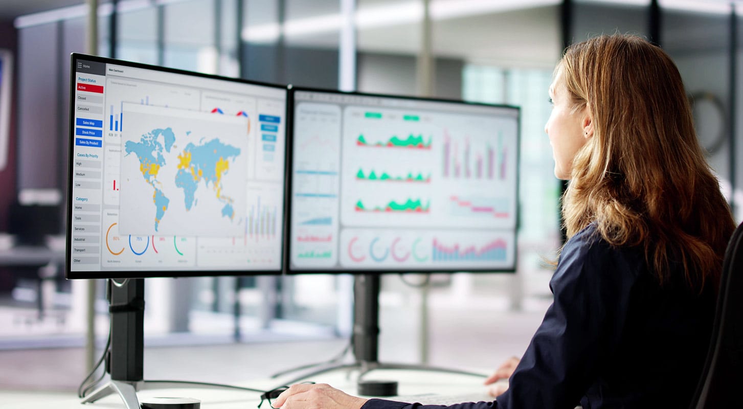 A woman at her desk with two computer screens, concentrating on data analysis.